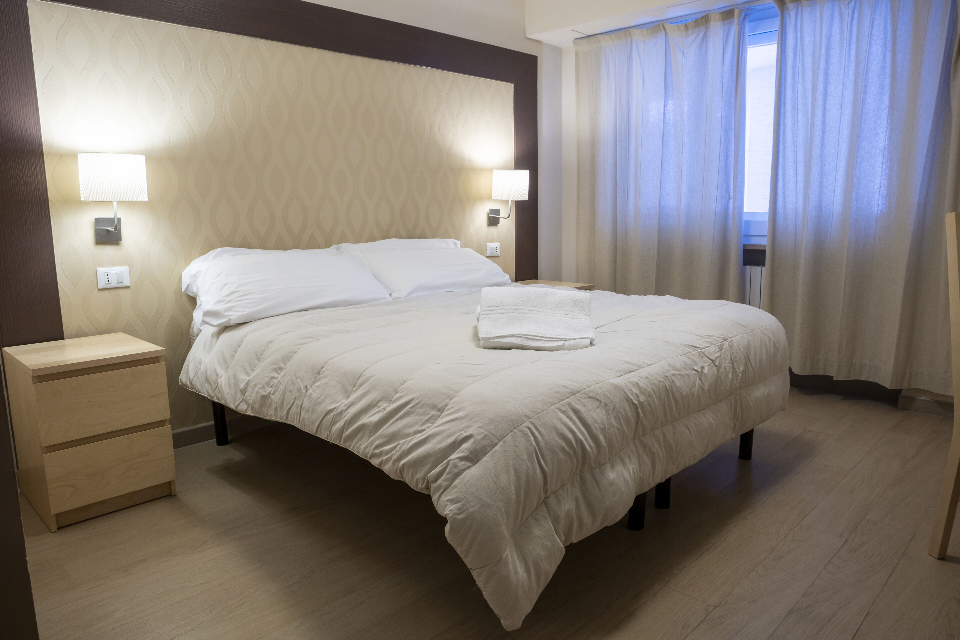 A neatly made bed with white bedding, a bedside table with drawers, and two wall-mounted lamps.