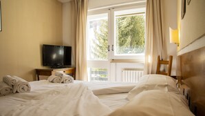 A hotel room with two beds, a television on a wooden nightstand, and a window with curtains. - Hotel Hermitage (Sestriere)