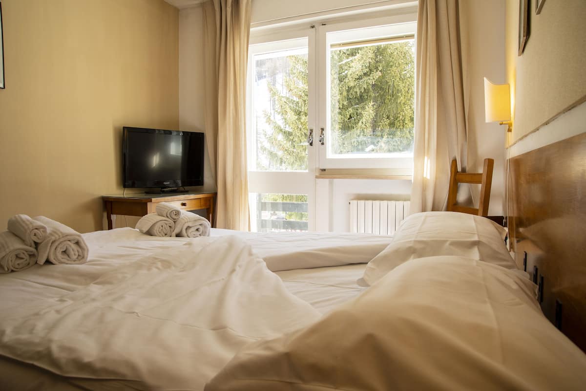 Une chambre d’hôtel avec deux lits, un téléviseur posé sur une table de chevet en bois et une fenêtre avec des rideaux.