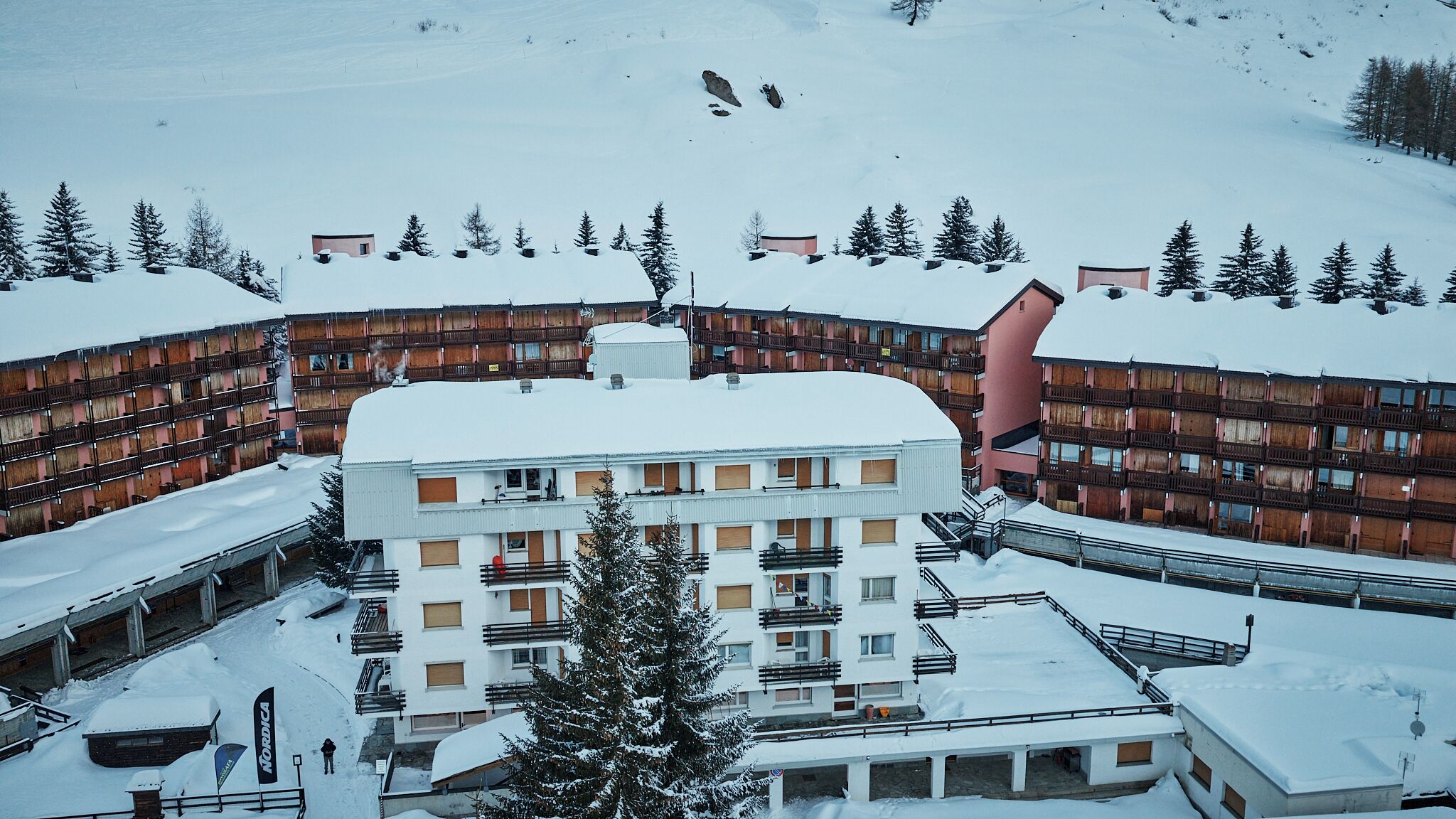 Estación de esquí cubierta de nieve con edificios de varios pisos, incluyendo un edificio central con balcones y un telesilla visible al fondo.