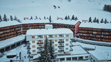Estación de esquí cubierta de nieve con edificios de varios pisos, incluyendo un edificio central con balcones y un telesilla visible al fondo.