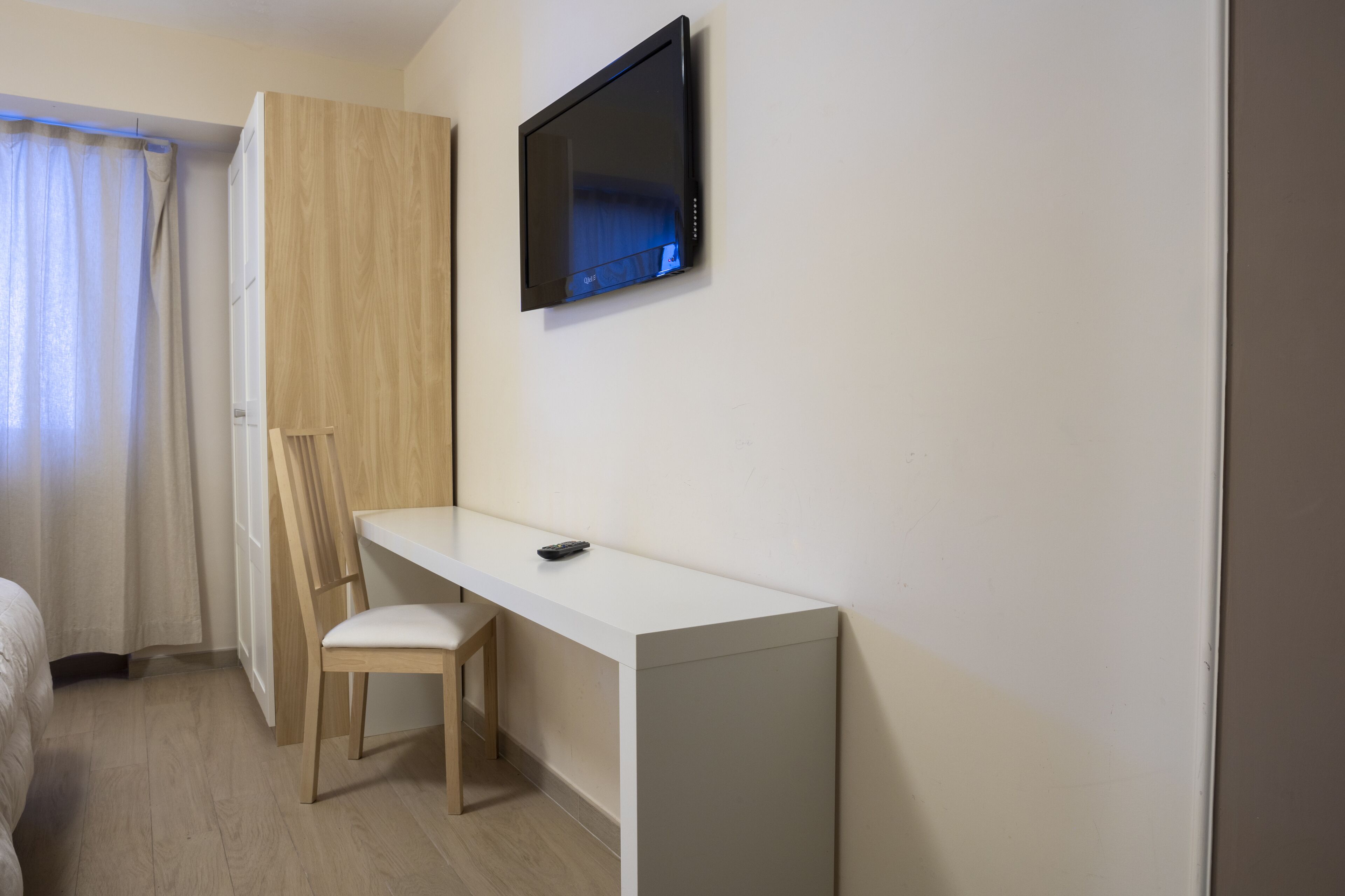 A hotel room with a white desk, a chair, a television mounted on the wall, and a window with curtains.