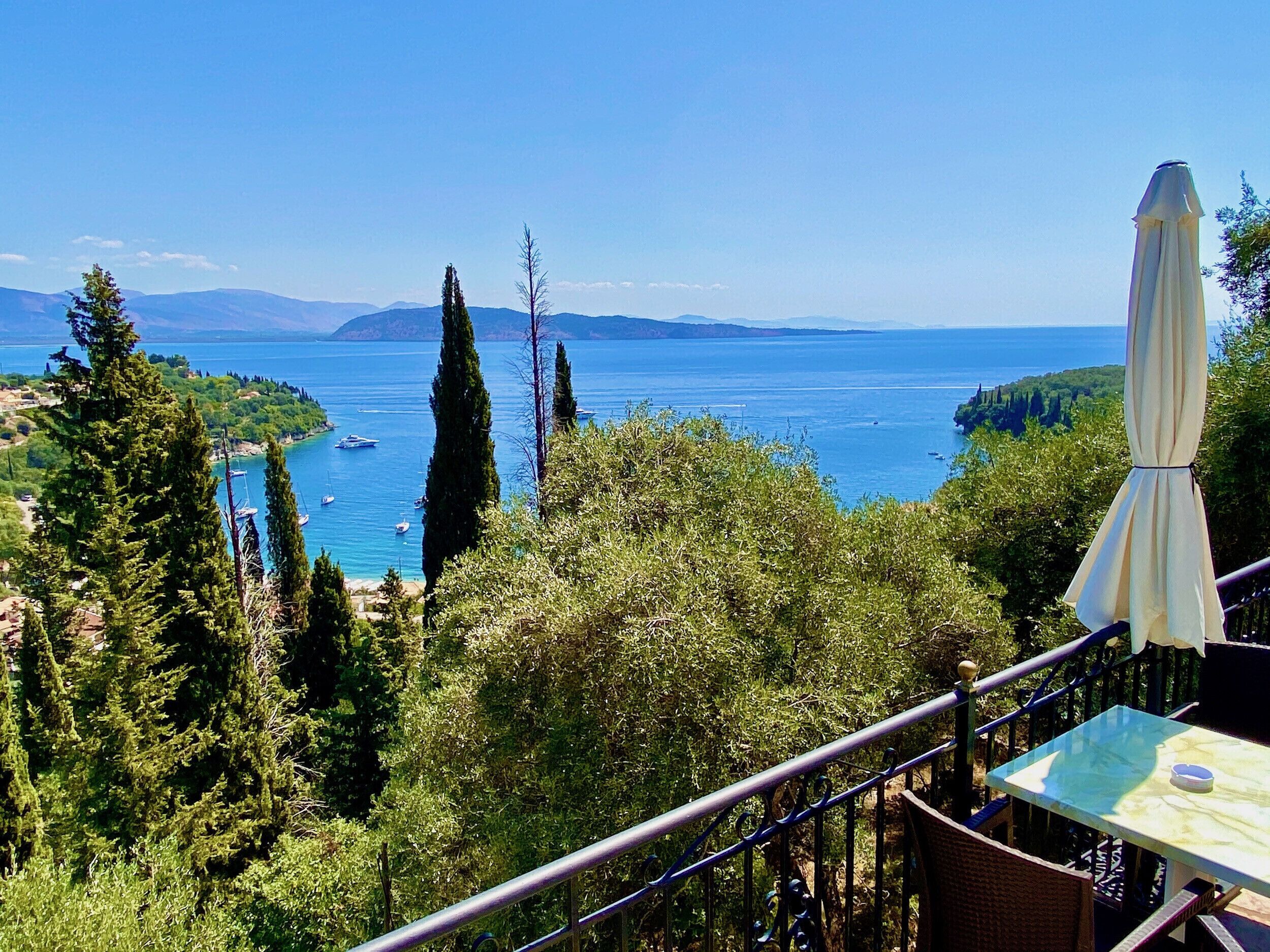 A terrace with a table and an umbrella overlooking a coastal landscape with boats and trees.