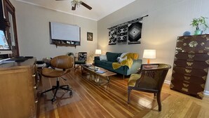 A living room with a wooden floor, a ceiling fan, a desk with a computer, a green sofa, and a decorative wall panel.