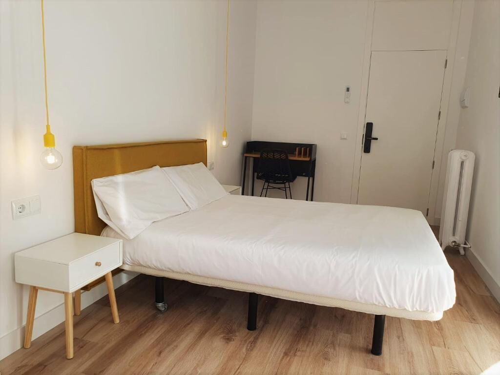 A single bed with a wooden headboard, a bedside table, a chair, and a desk with a chair.