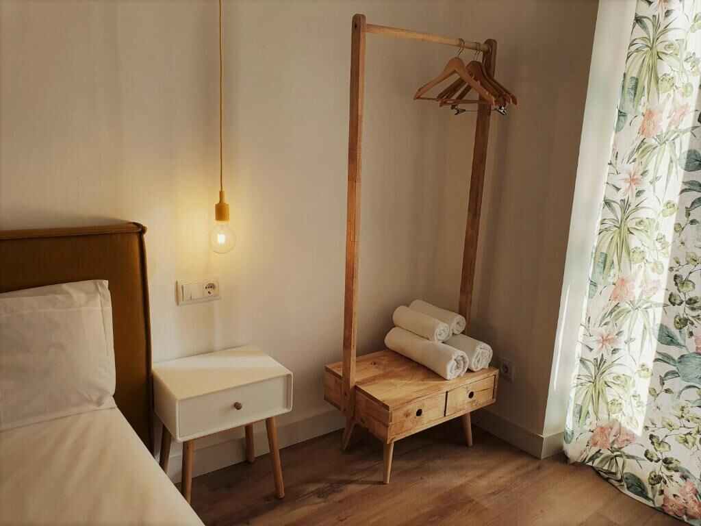 A room with a wooden bed, a bedside table with a lamp, a wooden hanging rack with towels, and a floral curtain.