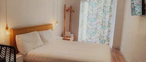 A bedroom with a wooden bed, a bedside table, a hanging light, a chair, and a window with floral curtains.