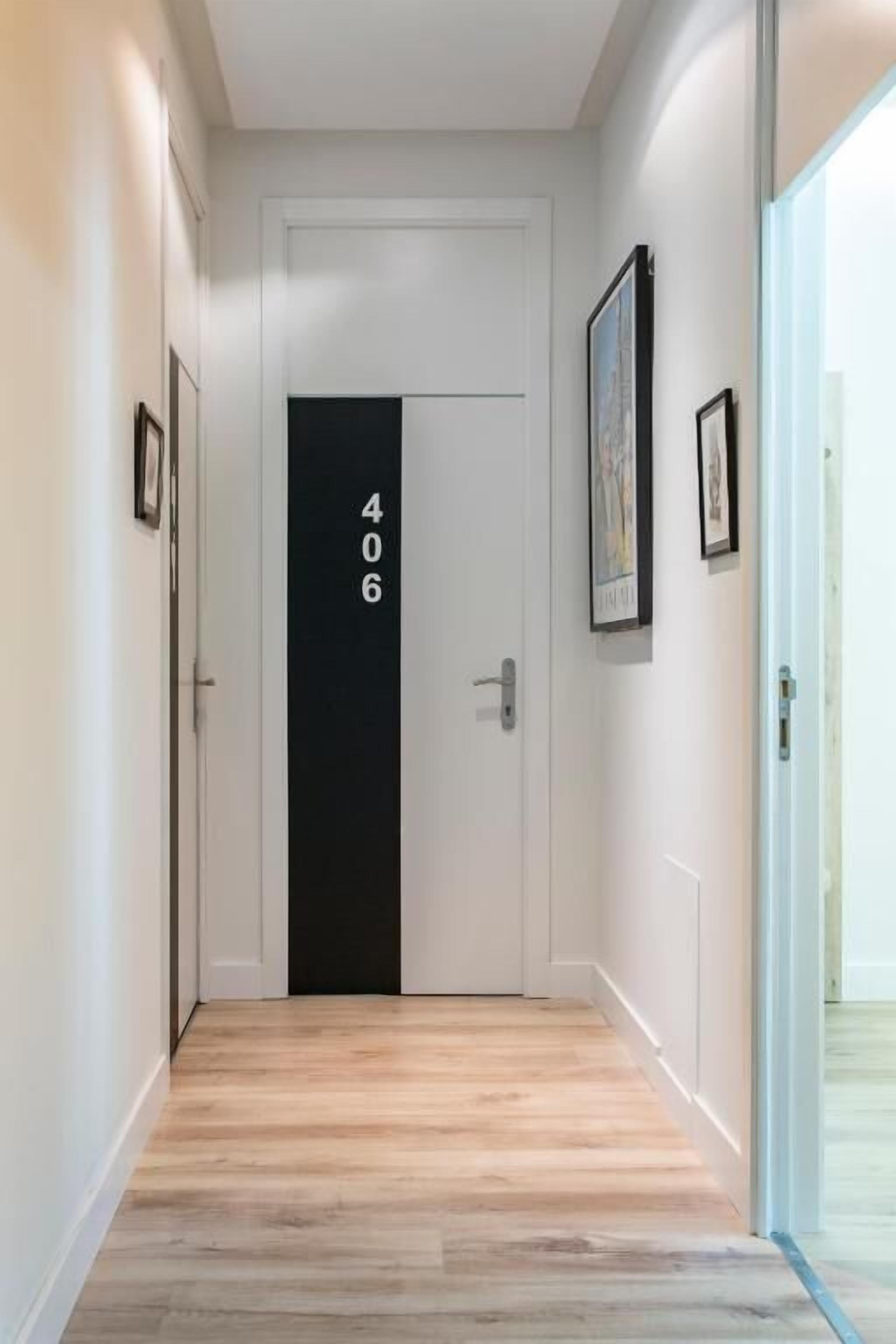 Un pasillo con una puerta blanca marcada con el número 406, suelo de madera e imágenes enmarcadas en las paredes.