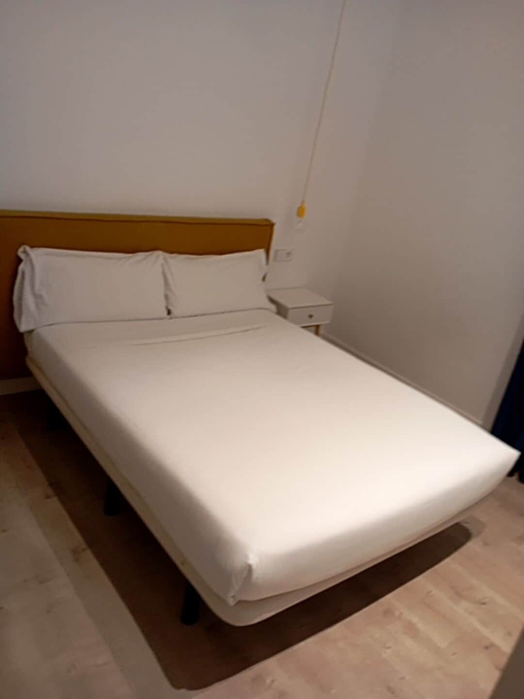 A single-bed room with a wooden headboard, white bedding, and a bedside table with a lamp.