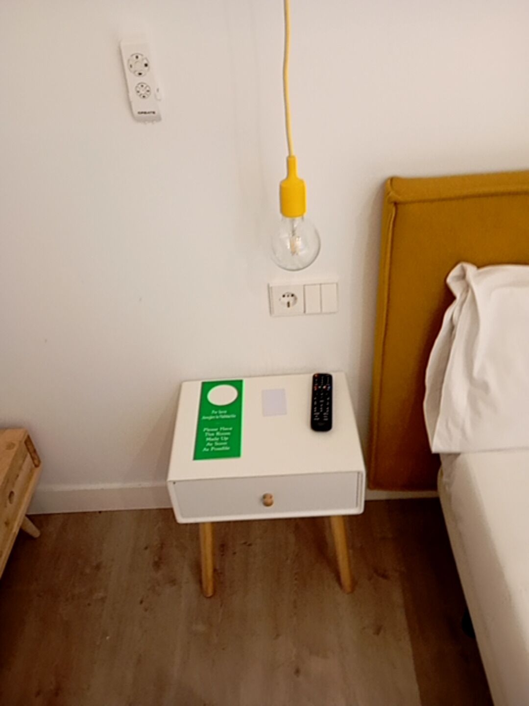 A small bedside table with a green sign, a remote control, and a yellow lamp plugged into a wall socket.