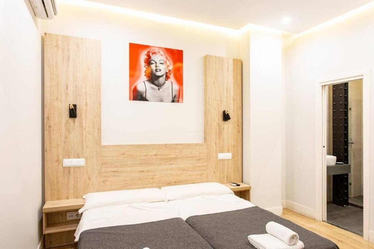 Een moderne hotelkamer met een houten hoofdbord, een bed met witte lakens, een grijze bank en een poster van een vrouw aan de muur.