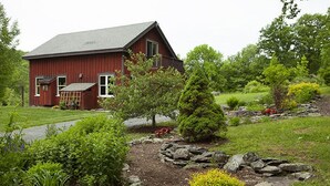 Une grange rouge au toit en pente, entourée d’un jardin bien entretenu où poussent divers arbustes et un petit arbre.