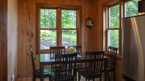 Une cuisine aux murs lambrés, avec une table et des chaises, un réfrigérateur et de grandes fenêtres donnant sur un paysage verdoyant.