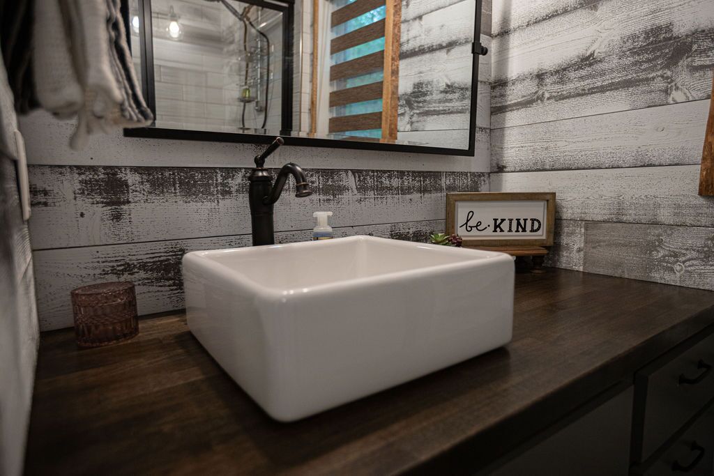 Un baño con lavamanos blanco, paneles de madera en la pared, un espejo y un cartel que dice 'be KIND'.