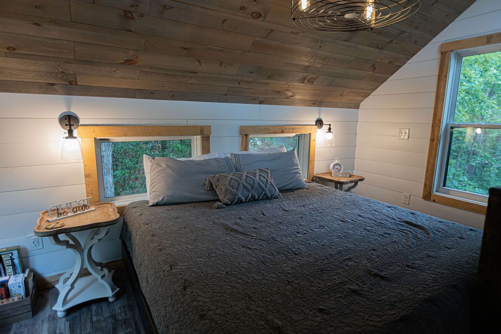 A bedroom with a wooden ceiling, a bed with a textured blanket, two bedside tables, and a window with a view of greenery.
