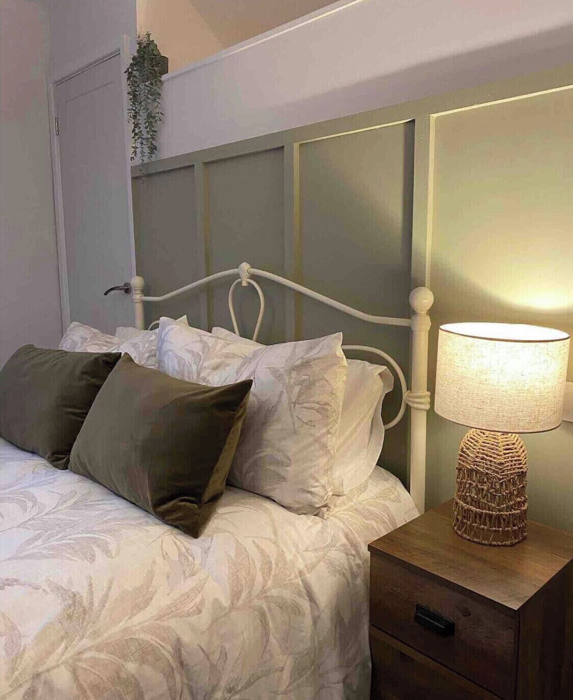 En seng med hvide sengetøj og to puder, et natbord med en lampe, og en plante hængt på væggen.