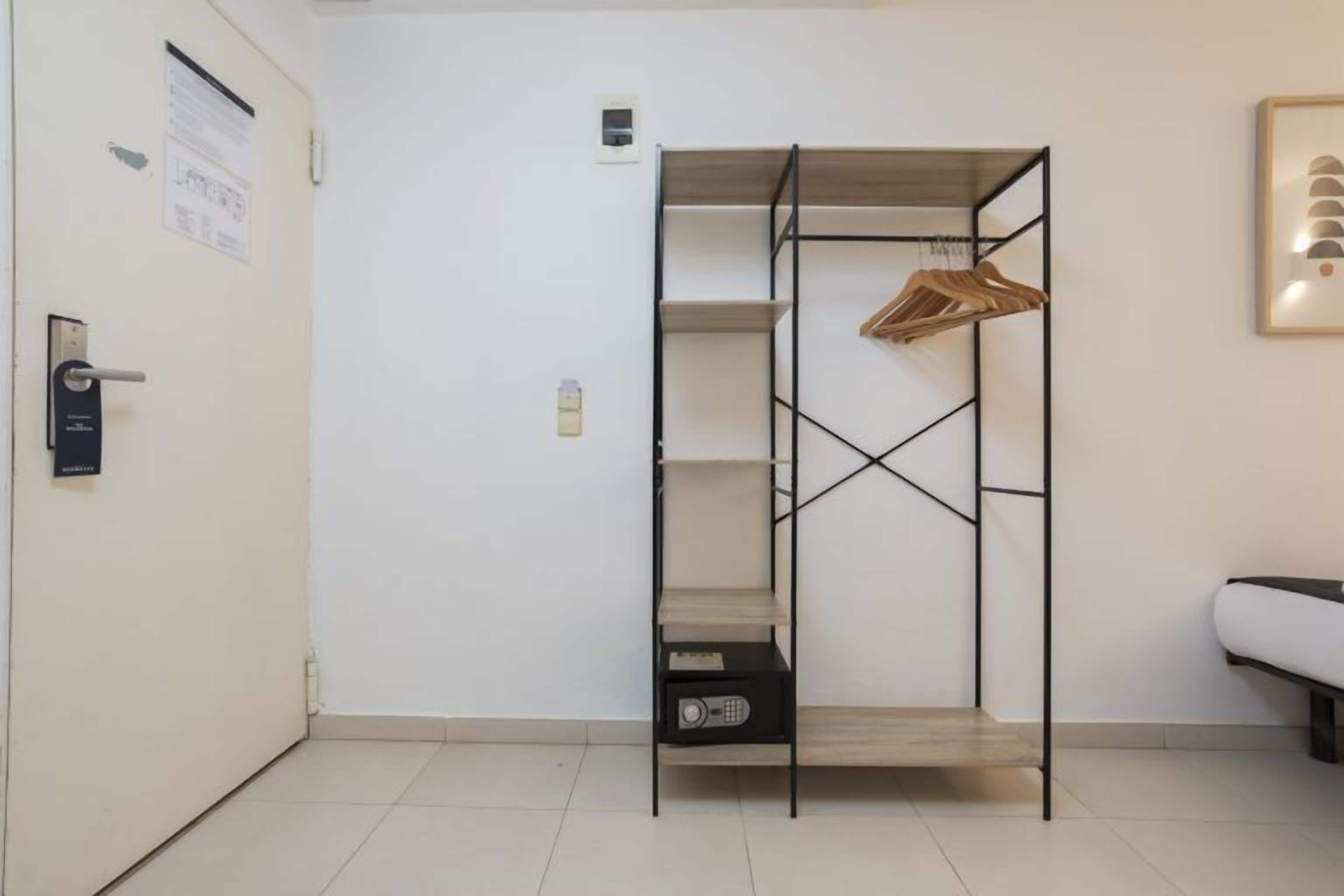 Una habitación con un estante metálico, una puerta con panel de seguridad, una mesita con un radio y un cuadro en la pared.