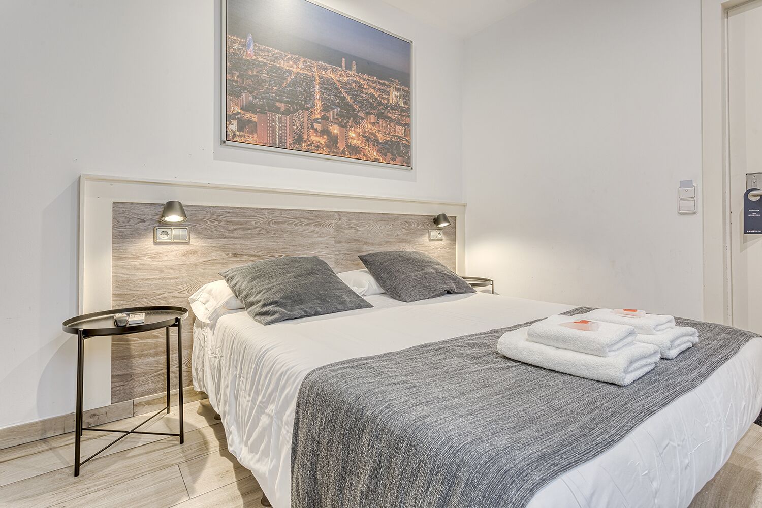 En pænt redt seng med et gråt tæppe og puder, et lille natbord med en lampe, og et bybillede over sengen.