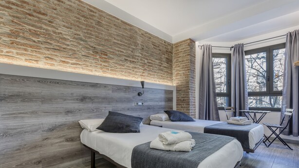 A hotel room with two beds, a brick wall, a wooden headboard, a window with curtains, and a small table with a chair. - Hostal Bedmates (Barcelona)
