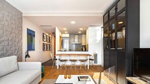 A modern living room with a sofa, a coffee table, and a TV. There is a dining area with a table and chairs. The kitchen is visible in the background with modern appliances. - Hostal Bedmates (Barcelona)