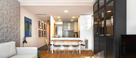Un salon moderne avec un canapé, une table basse et un téléviseur. On y trouve également un coin repas avec une table et des chaises. La cuisine, équipée d'appareils électroménagers modernes, est visible au fond.