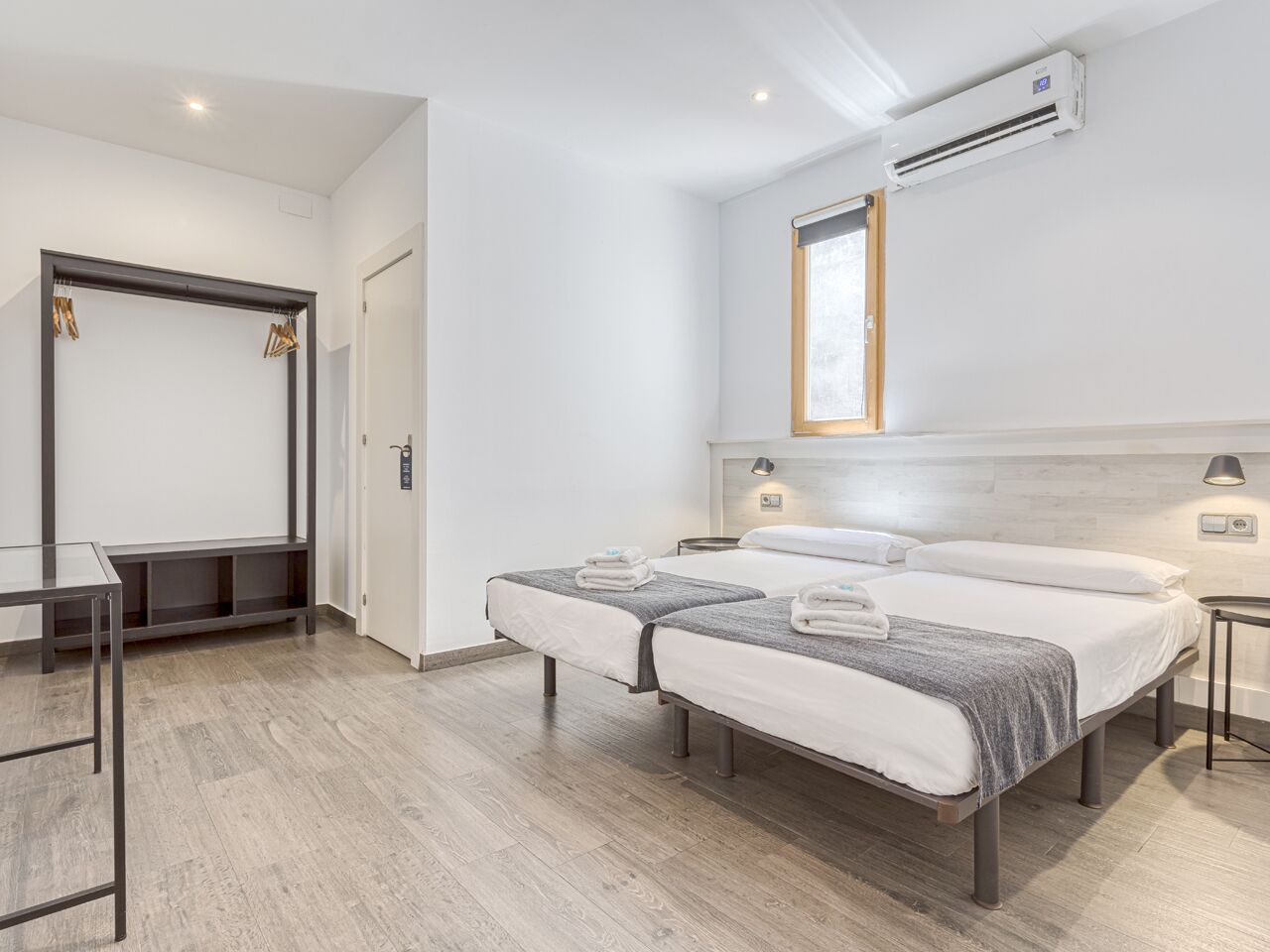 A modern hotel room with two beds, a desk, and a bench.