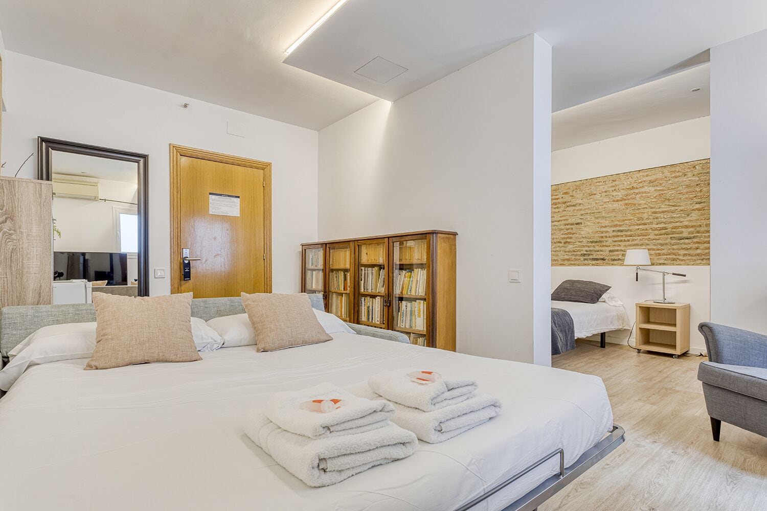 Habitación de hotel moderna con una cama grande, un armario de madera, una estantería y una zona de estar separada con una lámpara.