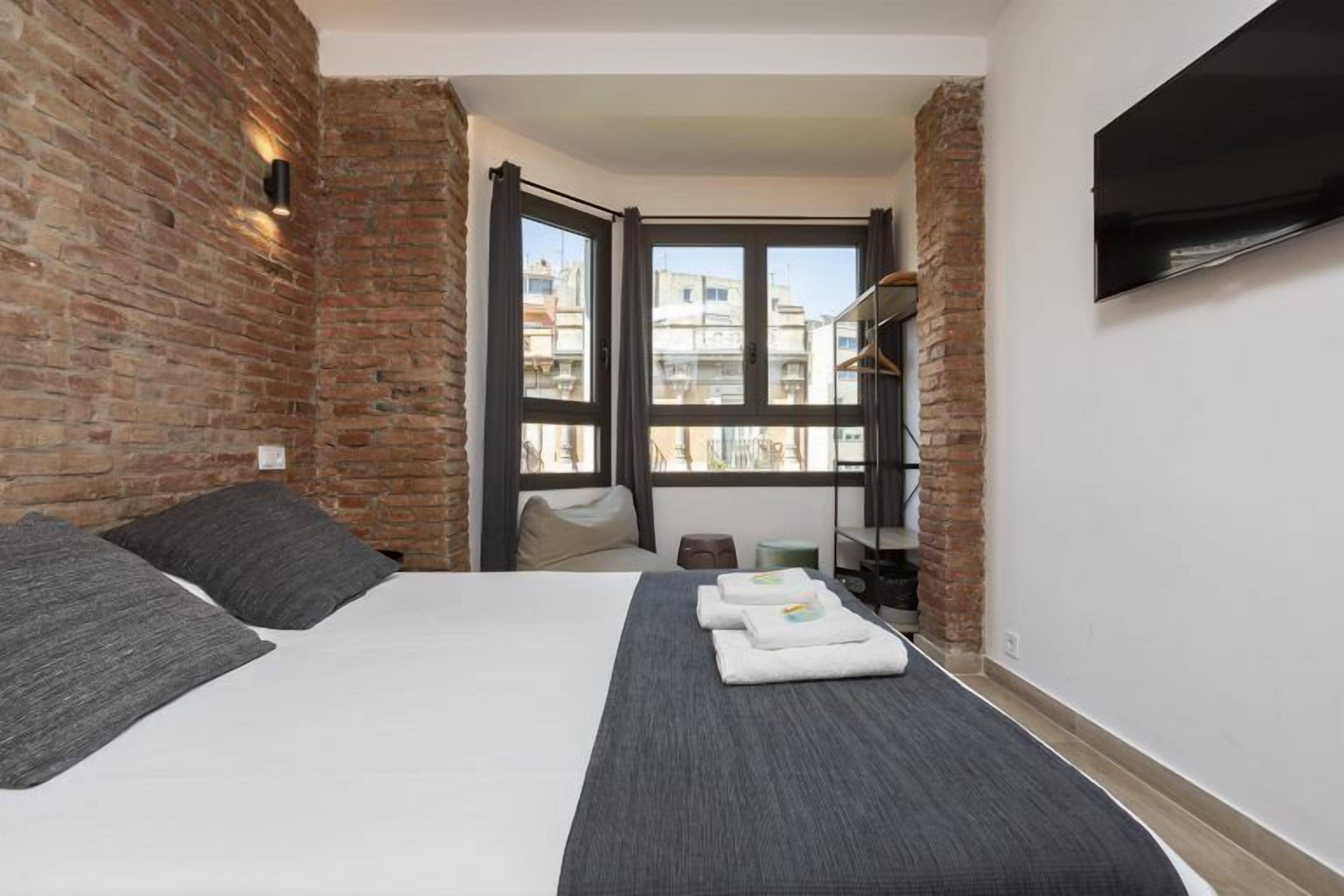 Quarto com parede de tijolos, cama com roupa de cama cinza, tapete cinza e janela com vista para edifícios.