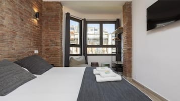 A bedroom with a brick wall, a bed with grey bedding, a grey rug, and a window with a view of buildings.