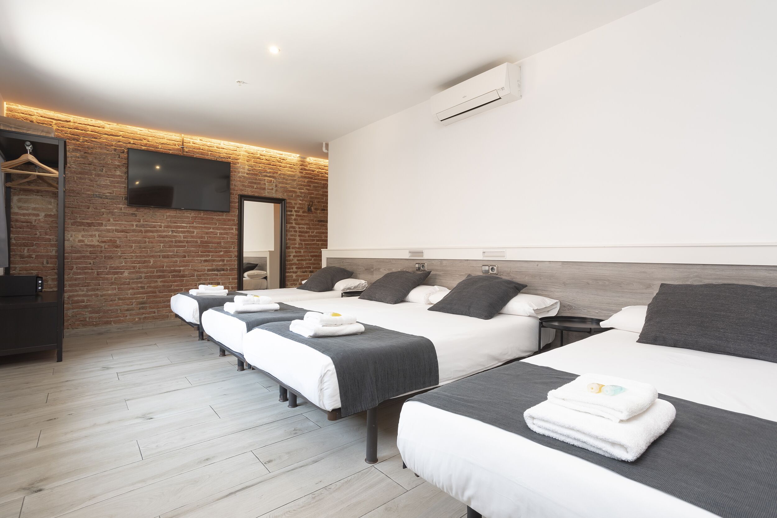A hotel room with three beds, a flat-screen TV mounted on a brick wall, and an air conditioning unit.