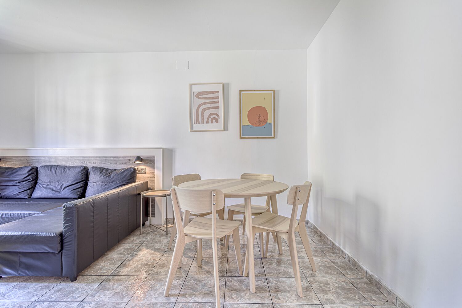 A modern living room with a grey sofa, a small table, and two chairs. There are two framed artworks on the wall.