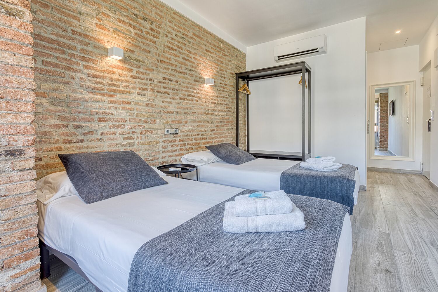 A hotel room with two beds, a brick wall, a large mirror, and a wooden floor.
