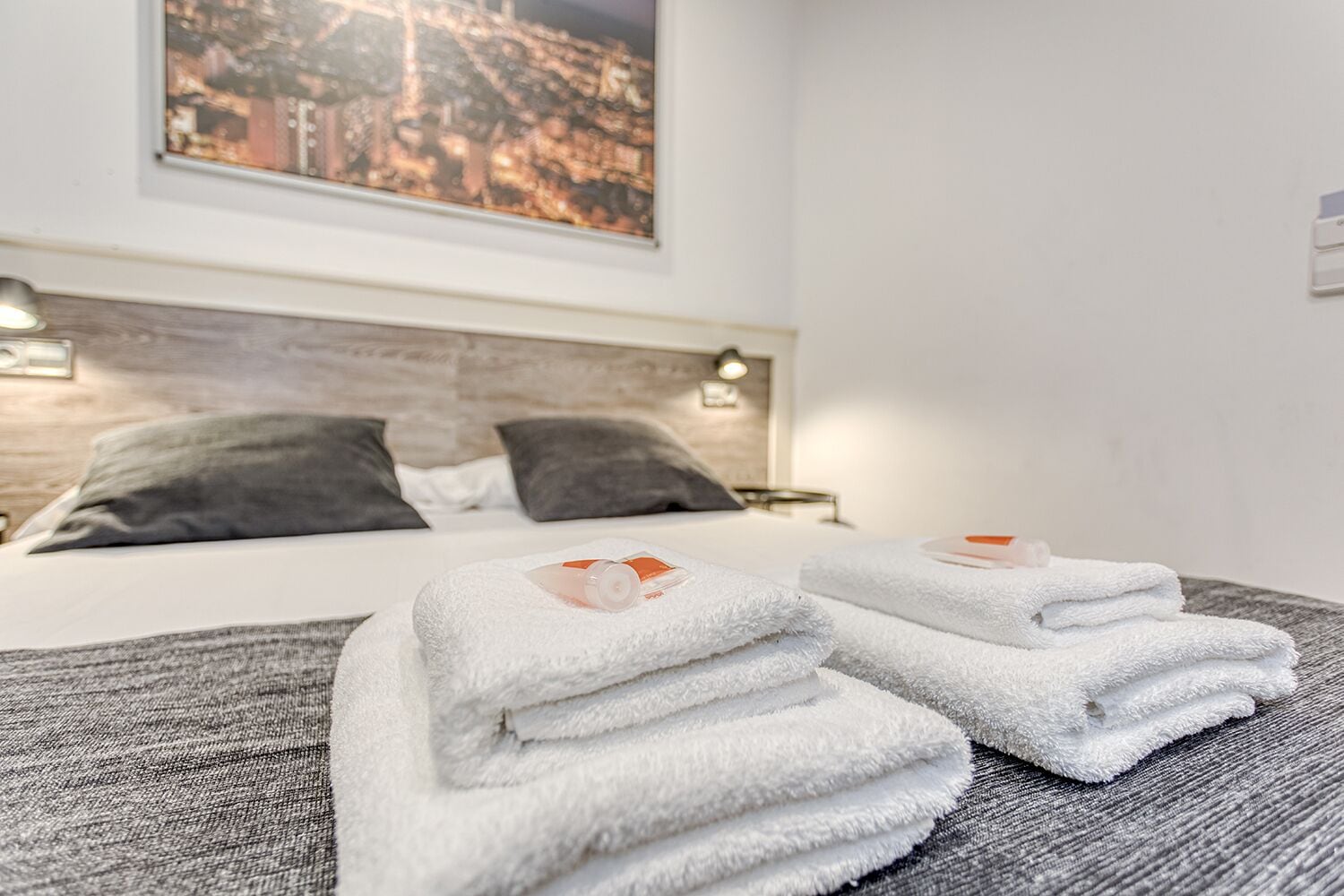 Una cama bien hecha con sábanas blancas y dos almohadas, vista panorámica de la ciudad a través de una ventana, y una lámpara fijada a la pared.