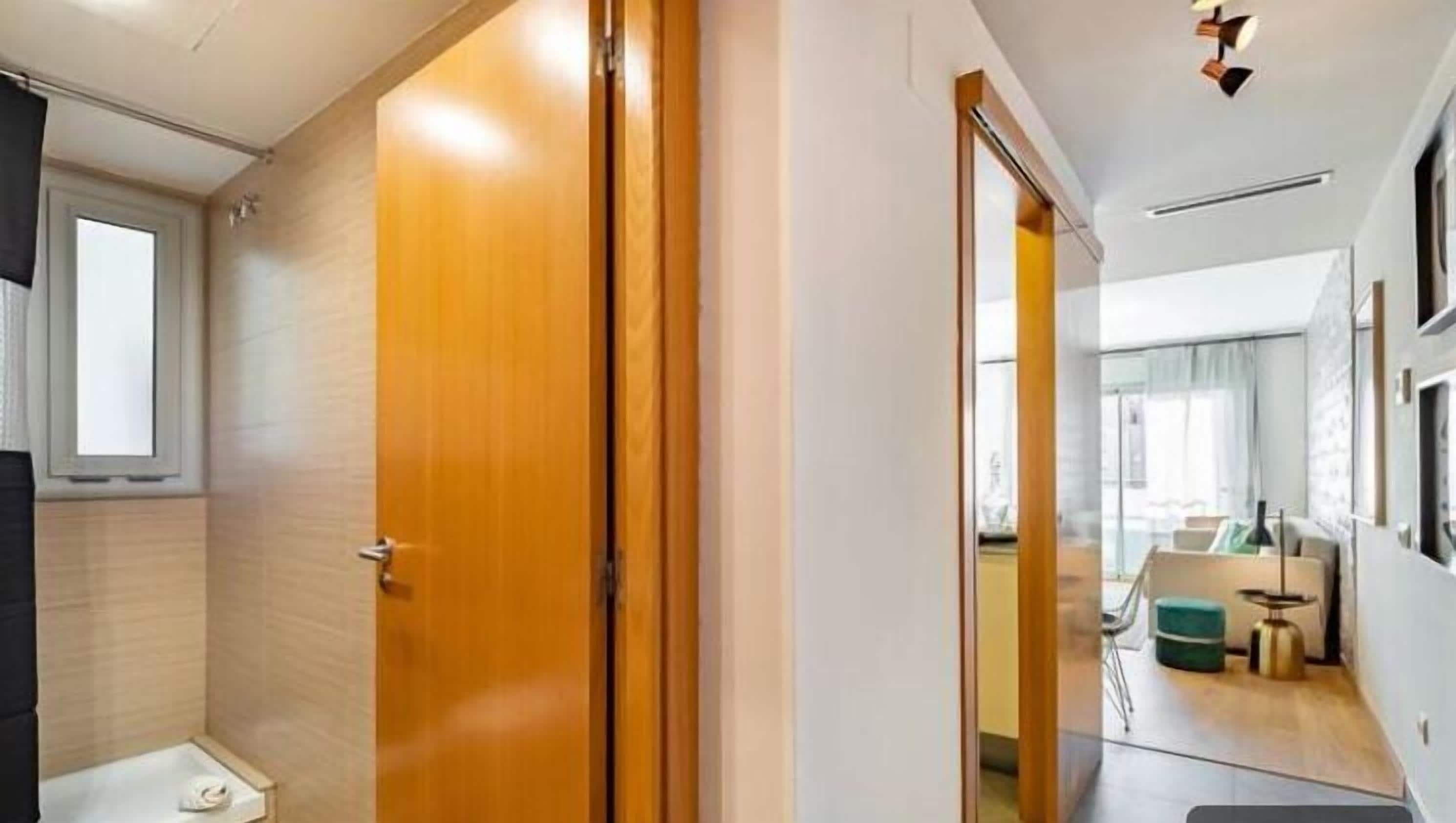 A modern hotel room with a wooden door, a small bathroom, and a view of the living area.