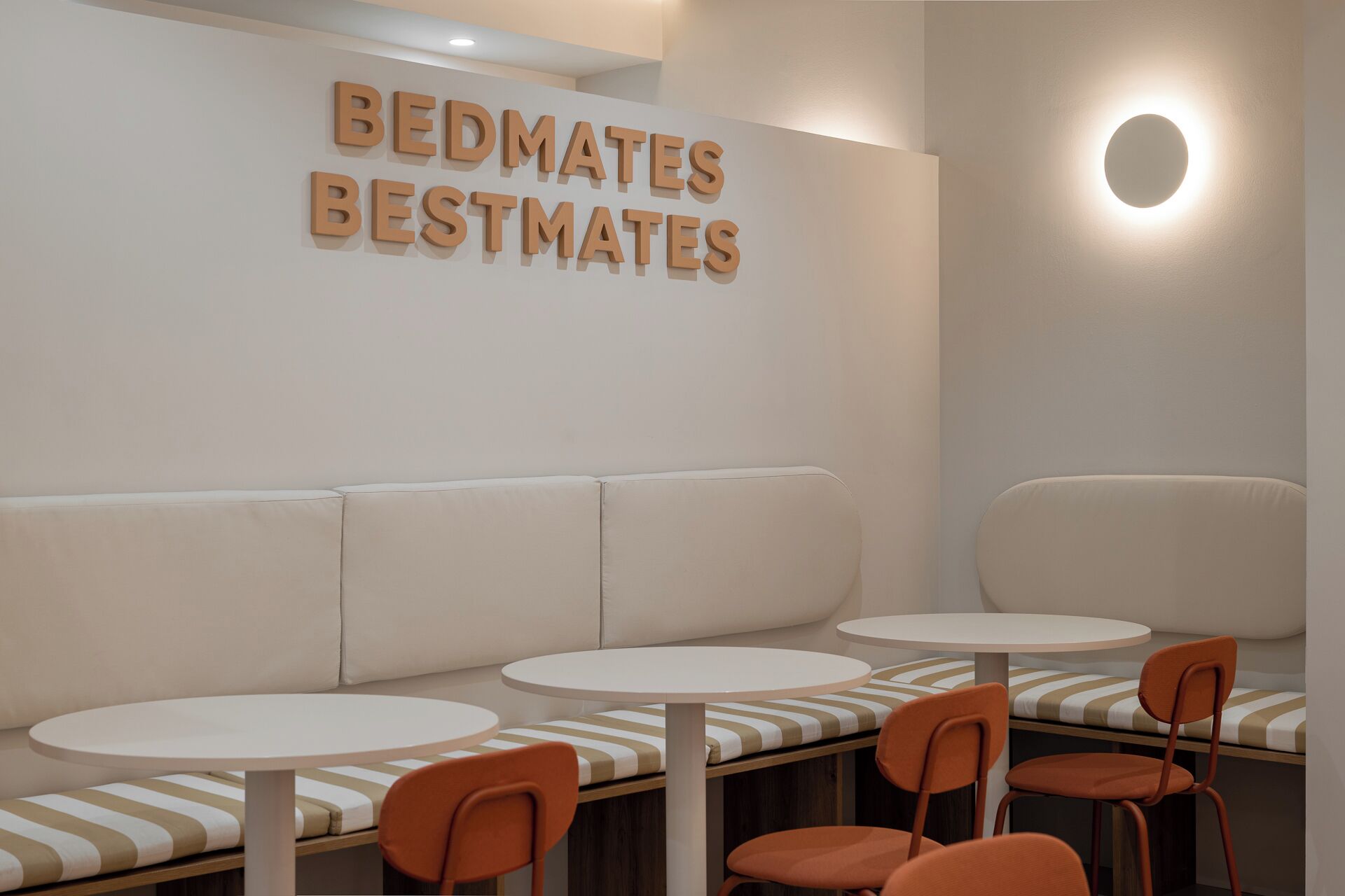 A seating area with a sofa and two round tables, each with a chair. The wall has a sign that reads 'BEDMATES BESTMATES'.