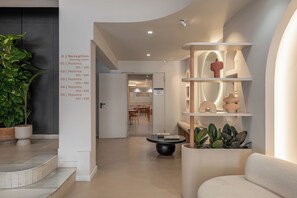 A modern hotel lobby with a reception desk, a dining area, and a seating area with a unique shelving unit and plants.