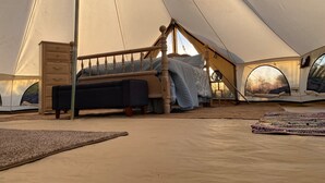 Een tent met een bed, een dressoir en uitzicht op de ondergaande zon door een rond raam.