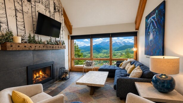 A cozy living room with a fireplace, comfortable seating, a large window with a mountain view, and a flat-screen TV mounted on the wall. - Mountain Views, Hot Tub & Walk to Summer Concerts (Crested Butte)
