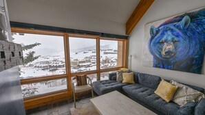 A cozy living room with a large window, a blue sofa, and a bear painting. - Mountain Views, Hot Tub & Walk to Summer Concerts (Crested Butte)