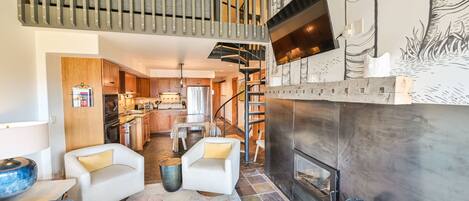 Amplia sala de estar con chimenea, una escalera y una cocina al fondo.