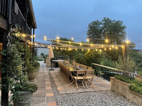 Un patio al aire libre con una larga mesa de madera y sillas, luces colgantes y una parrilla.