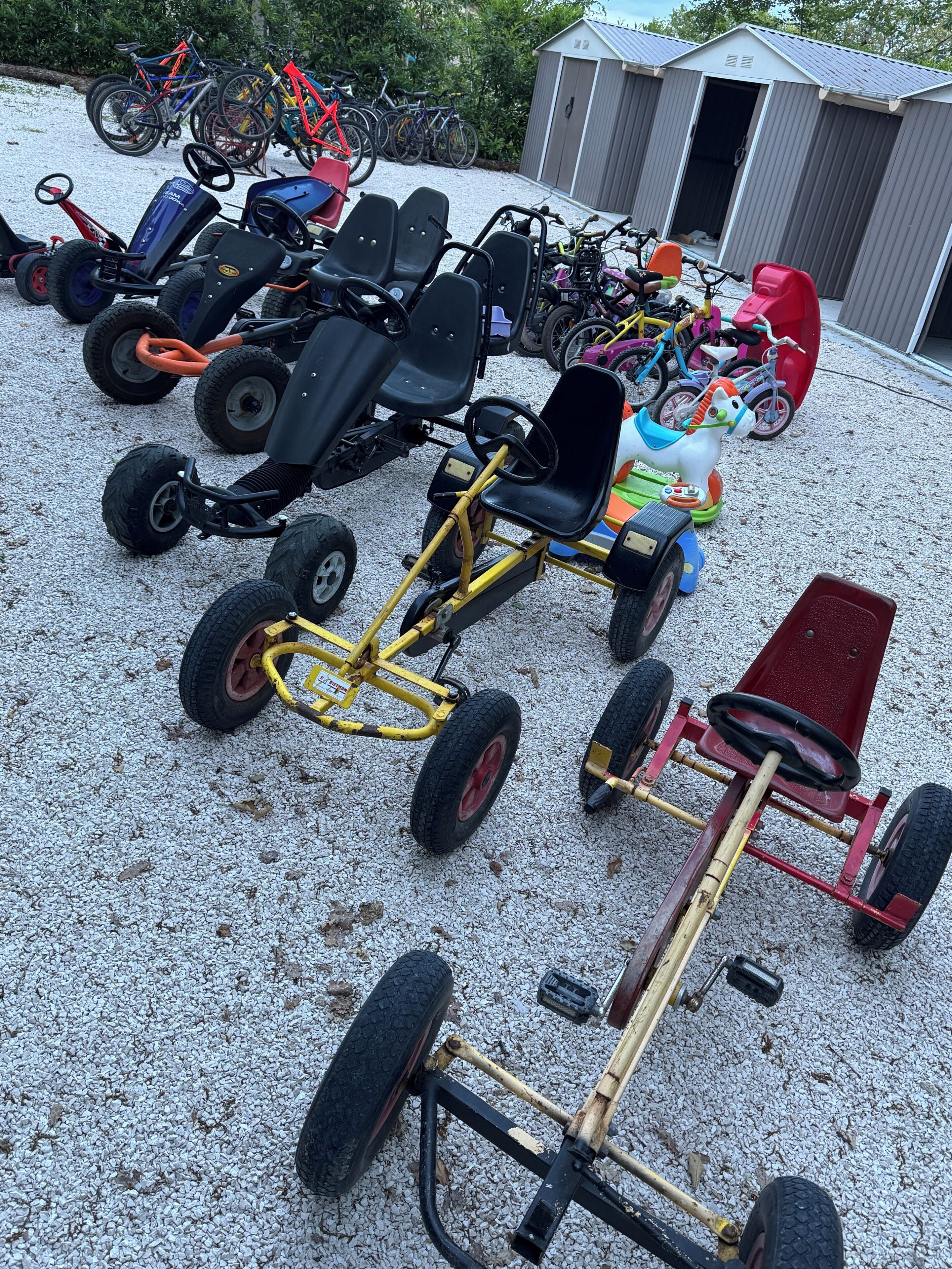 A collection of children's ride-on vehicles and bicycles parked on gravel.