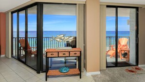 Una habitación con puerta de cristal corrediza que da a un balcón con vista al océano. Hay una pequeña mesa con un objeto decorativo y una silla visible en el balcón.