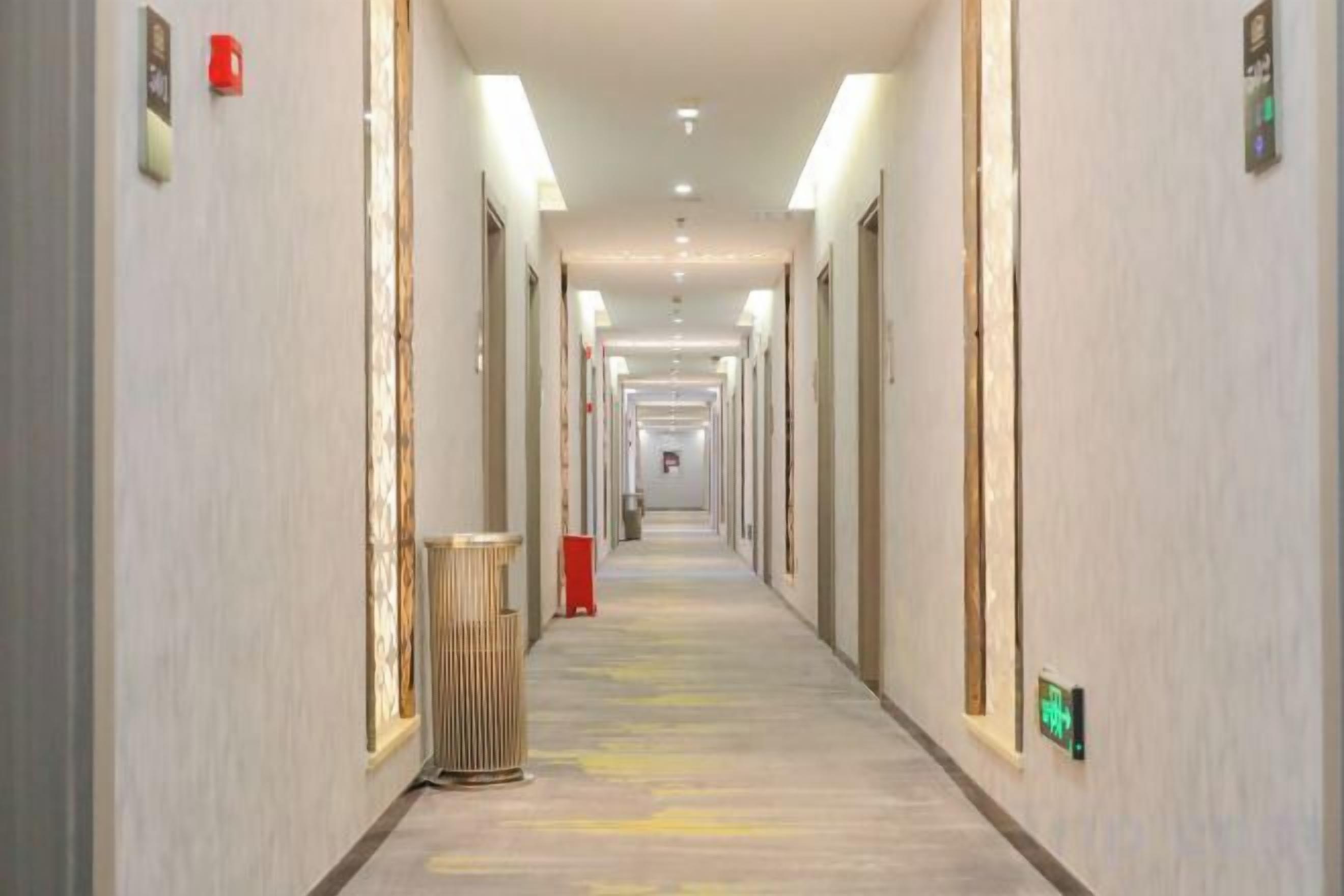 Un largo pasillo de hotel, estrecho, con puertas a ambos lados, un basurero y un objeto rojo a lo lejos.