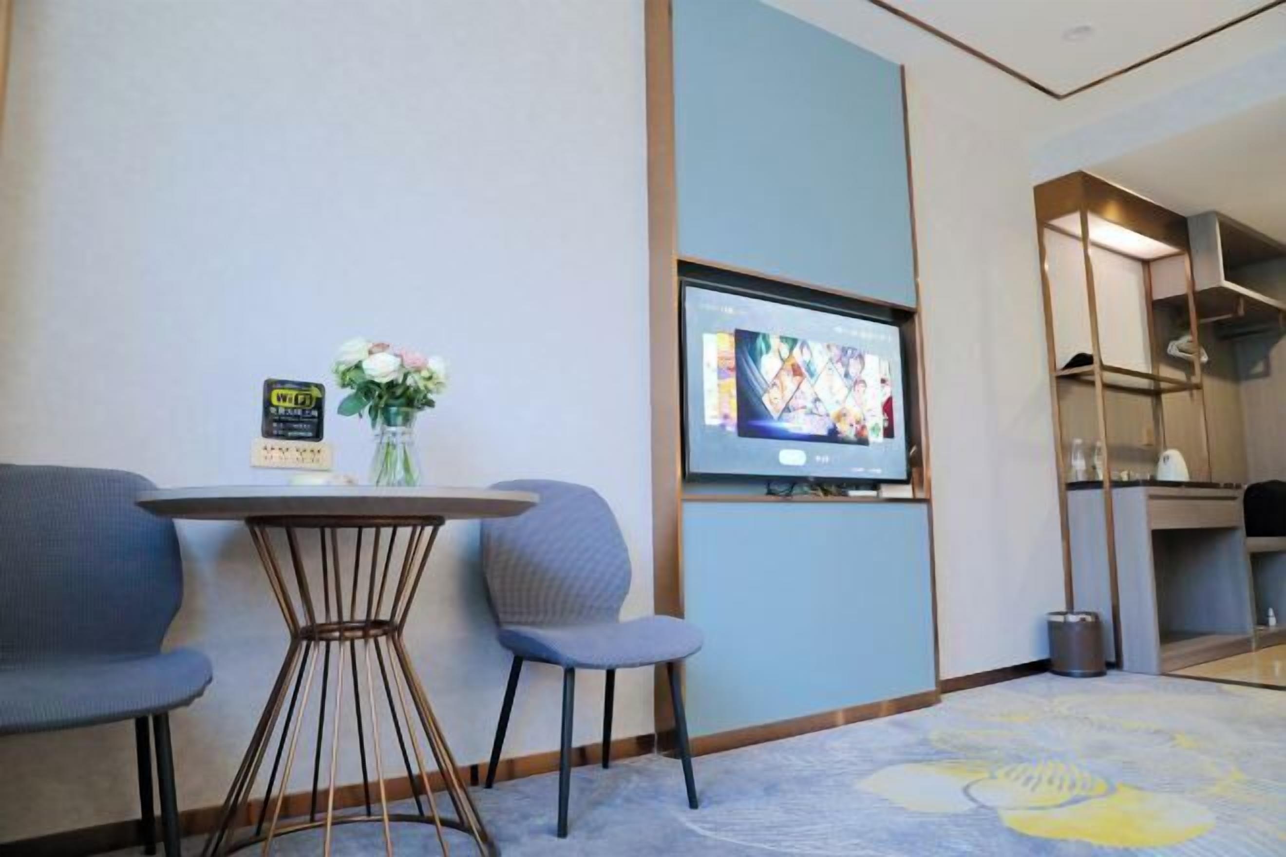 A hotel room with a TV mounted on the wall, a small round table with chairs, a vase with flowers, and a carpet with a floral pattern.