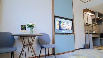 A hotel room with a TV mounted on the wall, a small round table with chairs, a vase with flowers, and a carpet with a floral pattern.