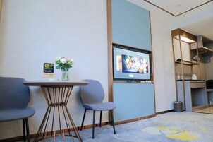 Habitación de hotel con televisor montado en la pared, una pequeña mesa redonda con sillas, un jarrón con flores y una alfombra estampada con motivos florales.