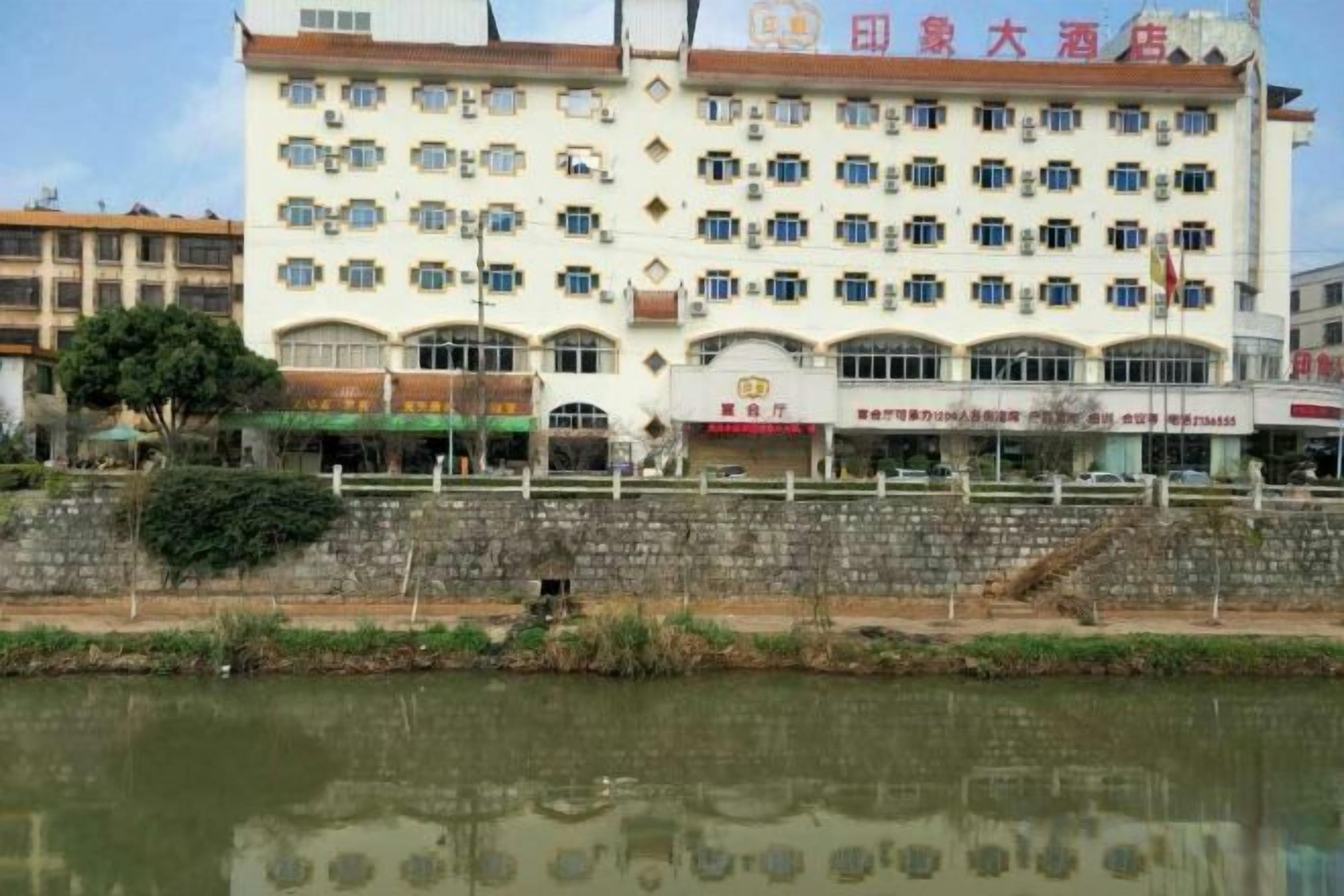 A multi-story hotel with a row of shops on the ground floor, a reflection in the water, and a stone wall in the foreground.