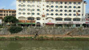 A multi-story hotel with a row of shops on the ground floor, a reflection in the water, and a stone wall in the foreground.