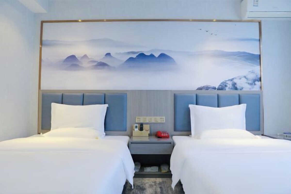 Une chambre d’hôtel avec deux lits, une grande œuvre d’art accrochée au mur, une table de chevet avec un téléphone et un objet rouge, et une tête de lit avec un panneau bleu clair.